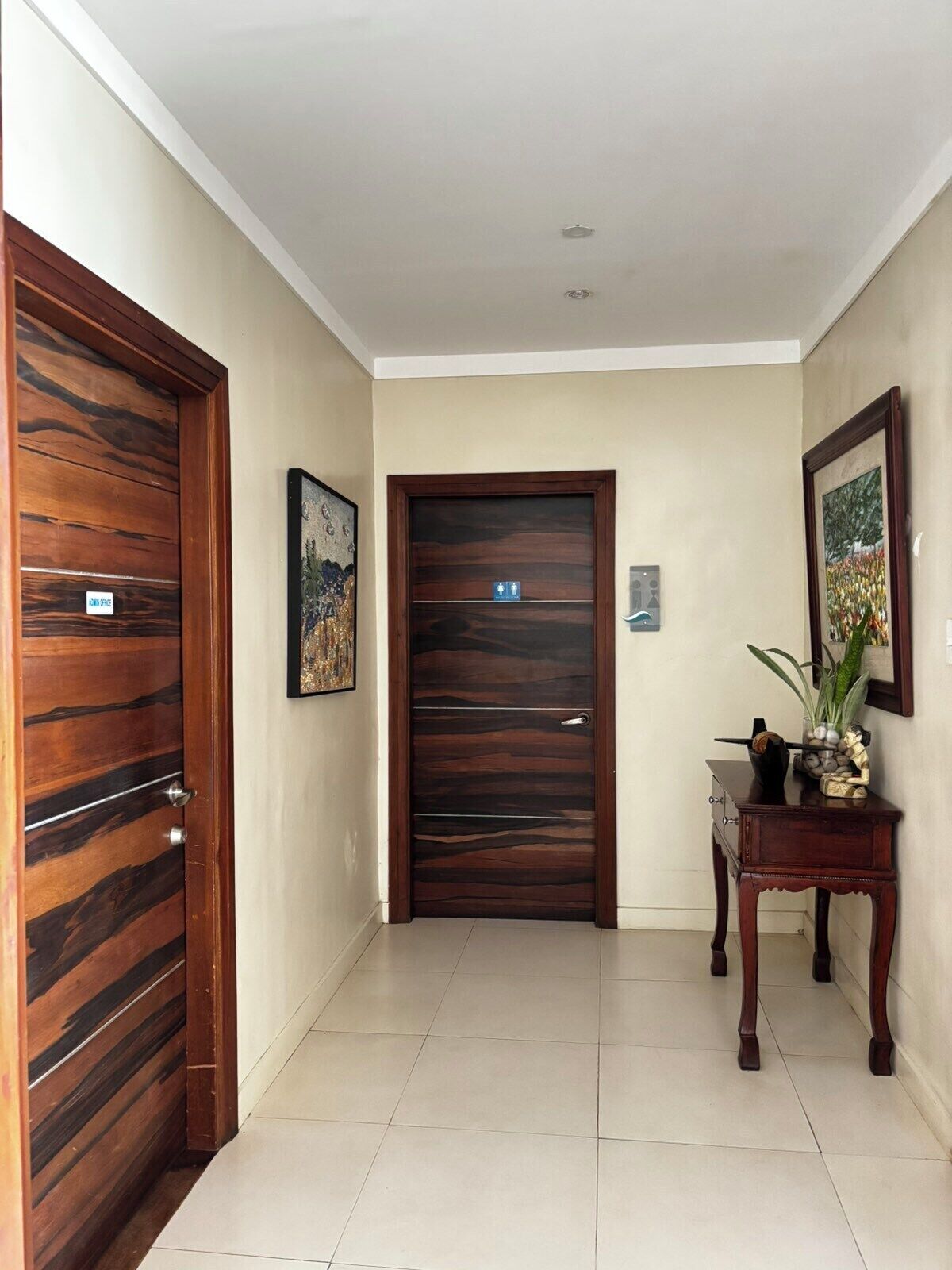 A hallway with a wooden door on the left, a framed picture on the wall, and a small table with a plant on the right.