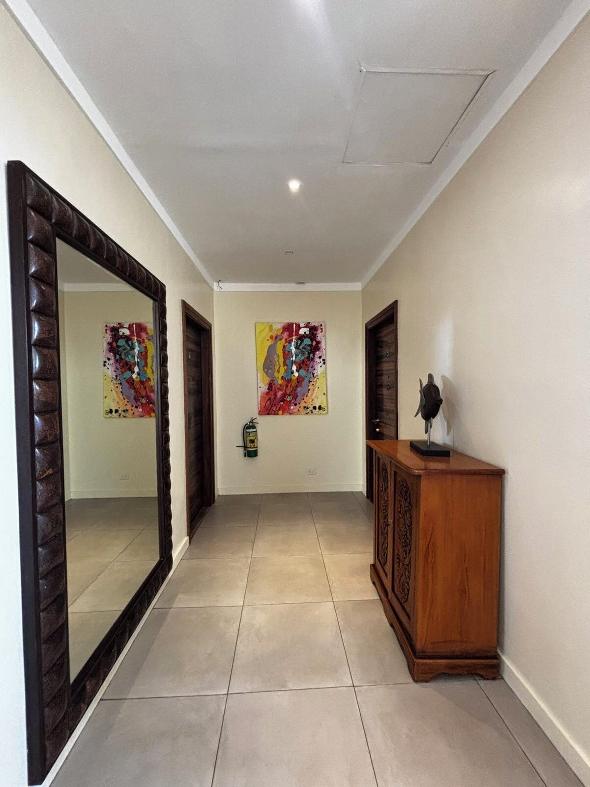 A hallway with a large mirror, two abstract paintings, a wooden cabinet, and a fire extinguisher.