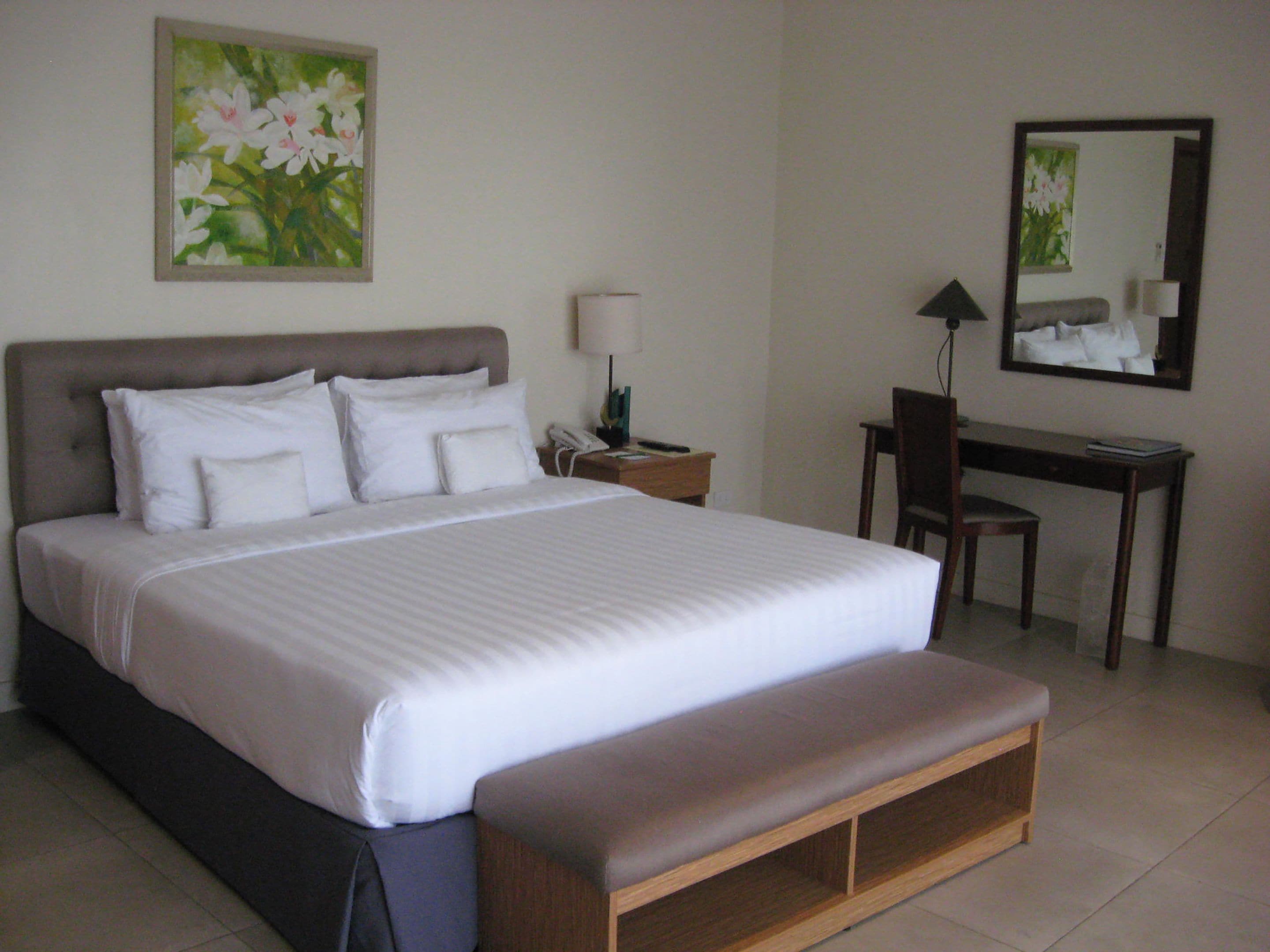 A neatly made bed with white linens, a bedside table with a lamp, a desk with a chair, and a painting on the wall.
