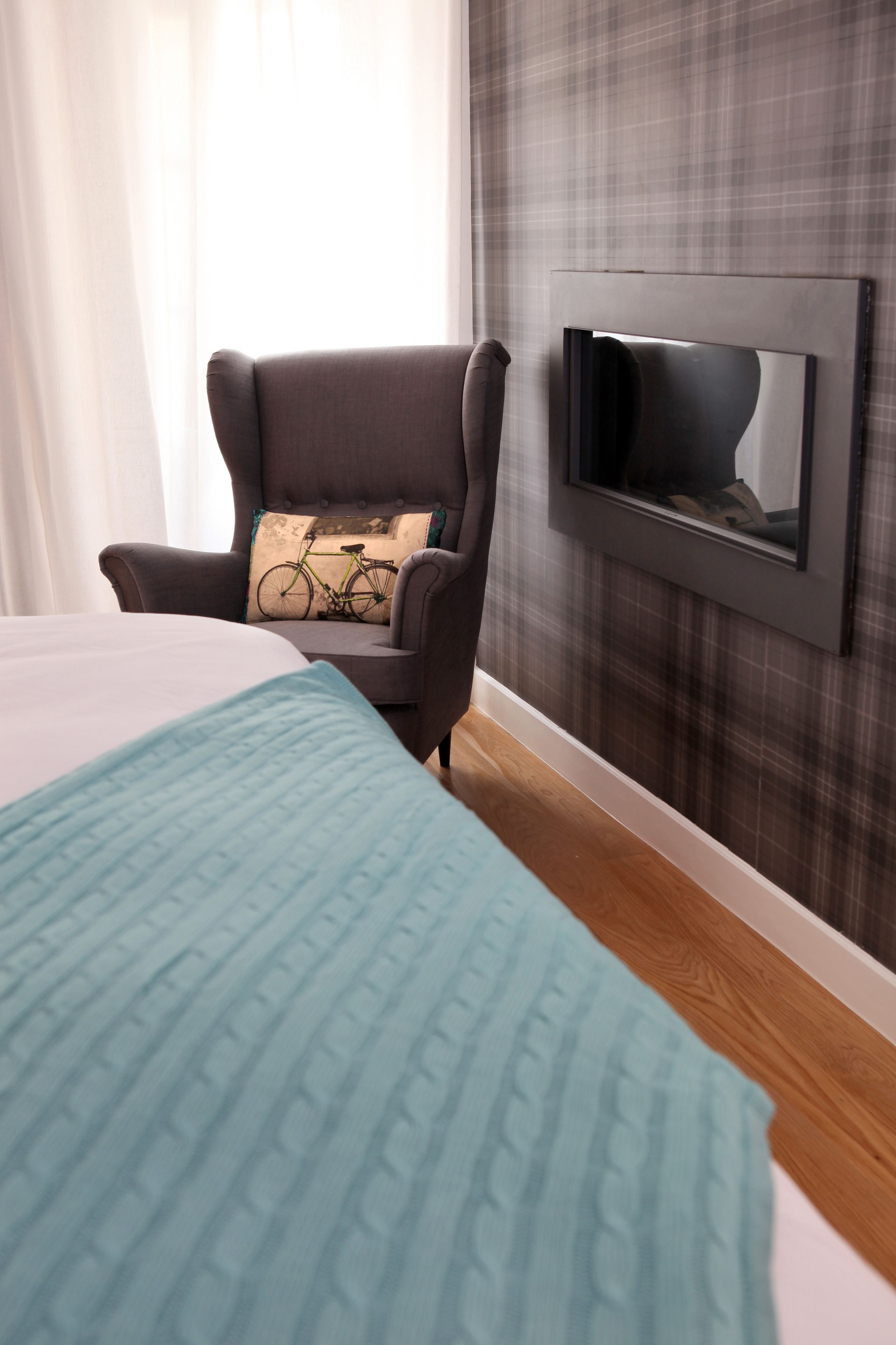 A hotel room with a bed, a chair, a television mounted on the wall, and a window with curtains.