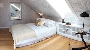 A bedroom with a sloped ceiling, a bed with white bedding, a bedside table with a lamp, and a chair.
