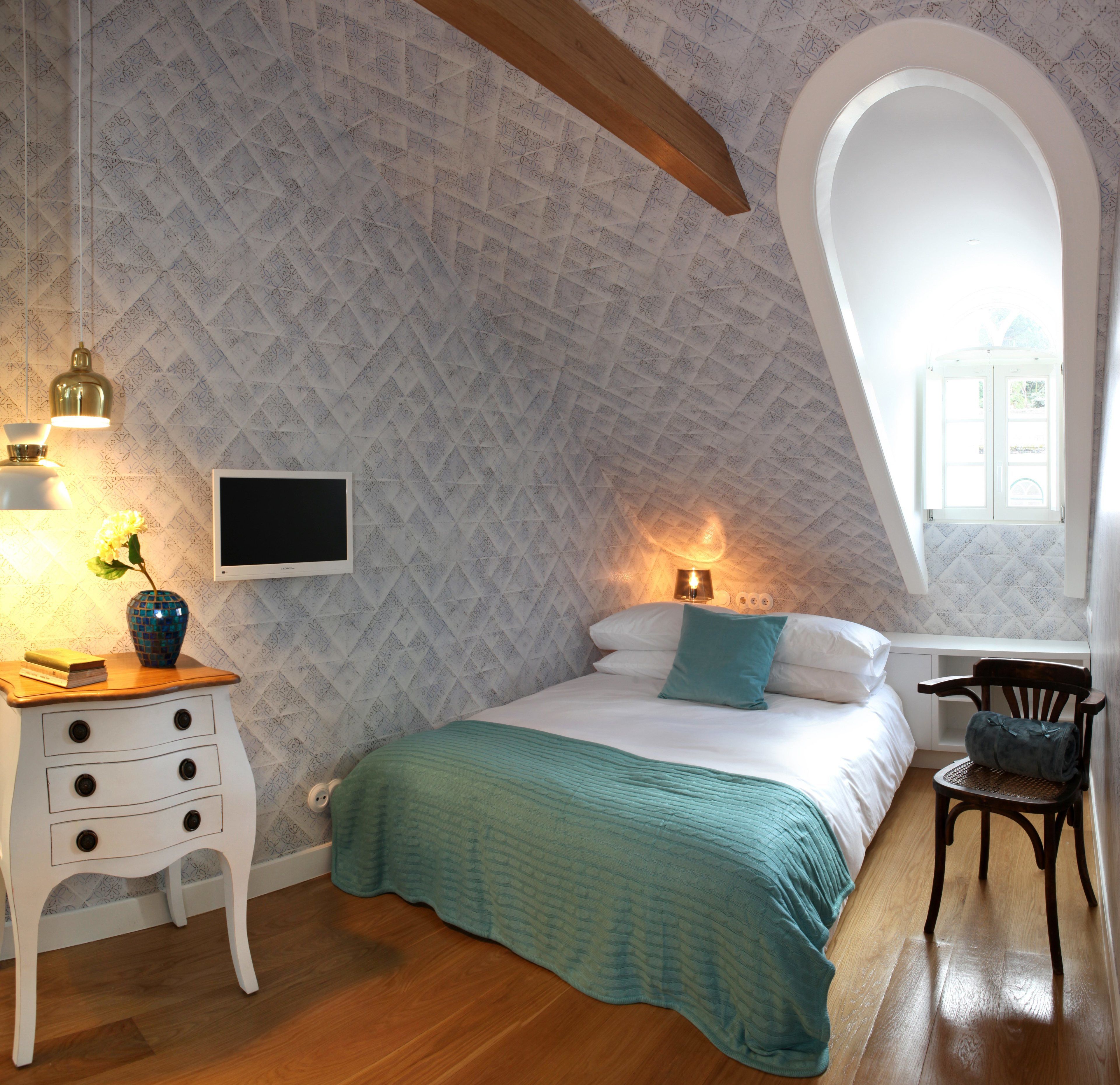 Ein Schlafzimmer mit geneigter Decke, einem Bett mit aquamarinblauer Decke, einem Nachttisch aus Holz mit einer Vase und Büchern, einem Sessel und einem an der Wand befestigten Fernseher.