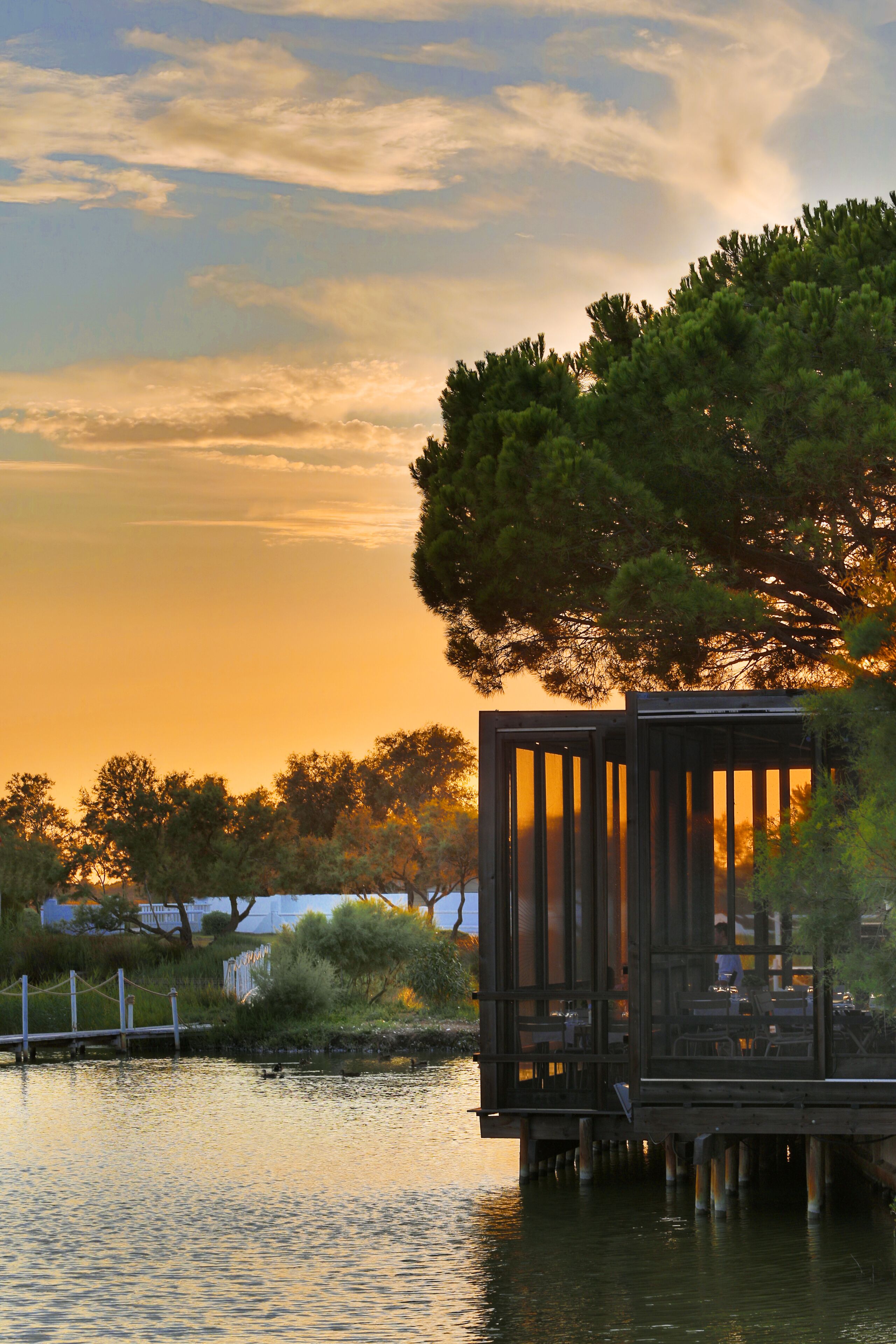 A wooden structure with a glass enclosure overlooking a body of water, surrounded by trees and a sunset sky.