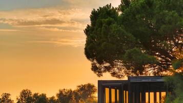 A wooden structure with a glass enclosure overlooking a body of water, surrounded by trees and a sunset sky.