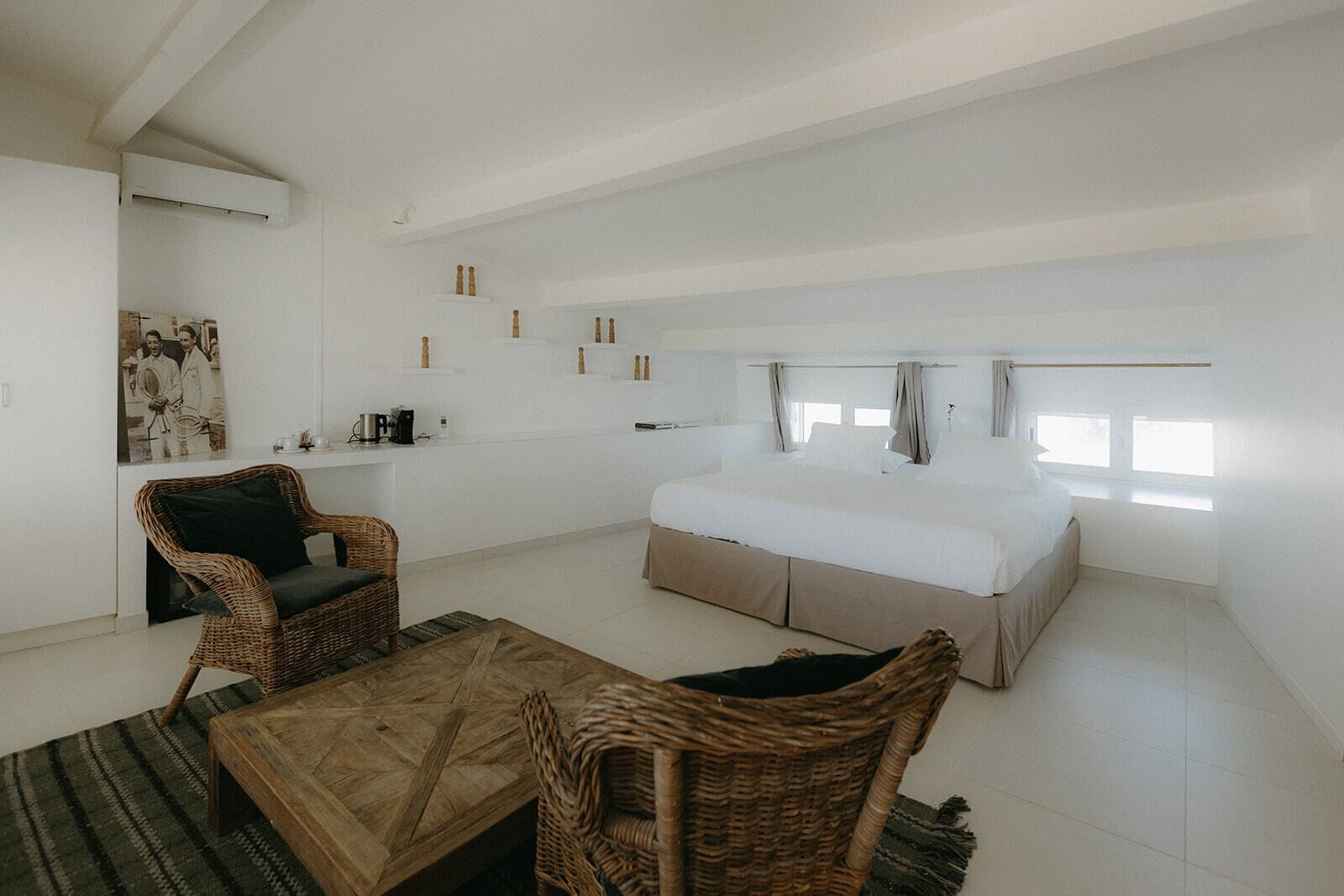 Een ruime kamer met een groot bed, twee rieten fauteuils, een houten bijzettafeltje en een klein nachtkastje.