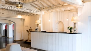 A reception area with a wooden reception desk, two lamps, a vase with flowers, and a seating area with cushioned chairs. - Mas de la Fouque Hôtel & Spa (Saintes-Maries-de-la-Mer)
