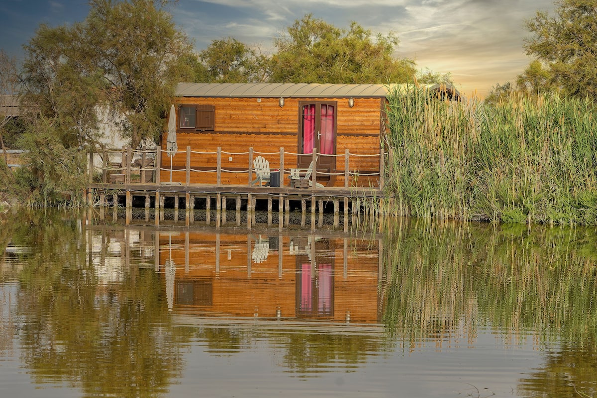 A wooden cabin with a deck, a chair, and a table by a calm body of water.