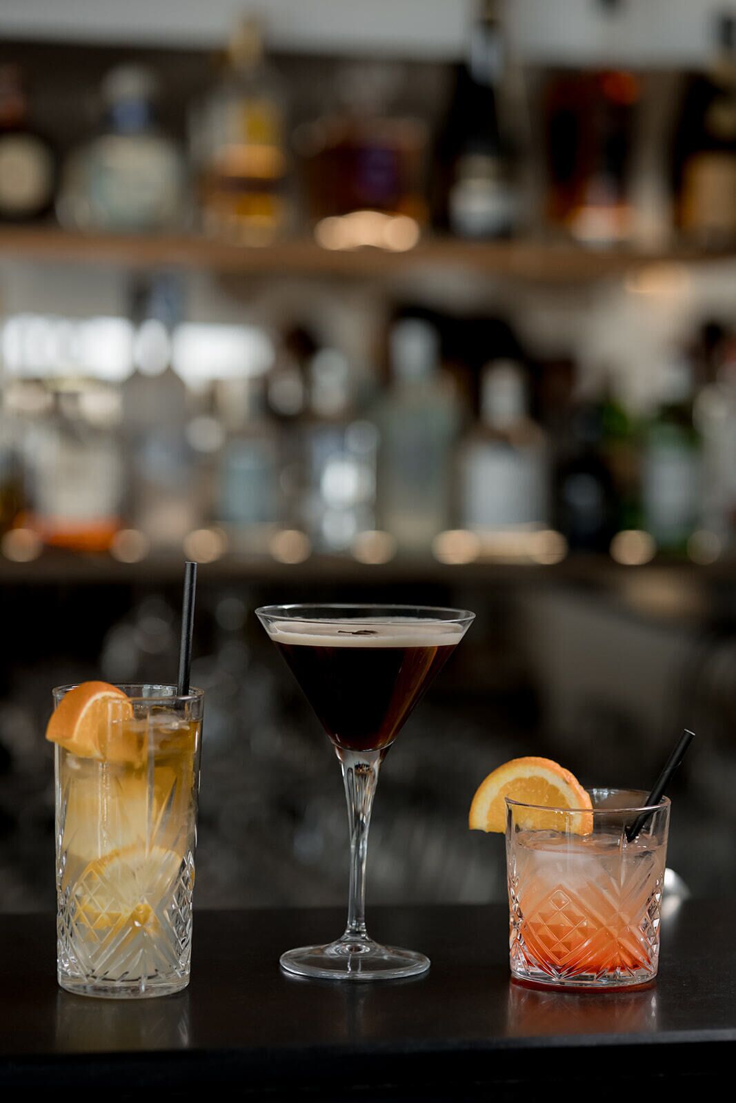 Tre cocktail su un bancone bar, con sullo sfondo sfocato scaffali.