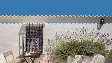 A patio with a white wall, a window with blue patterned shutters, a table with chairs, and a planter with lavender plants.