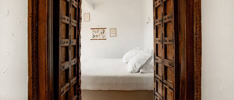 Un pasillo estrecho que lleva a un dormitorio con cama, paredes blancas y puertas de madera.
