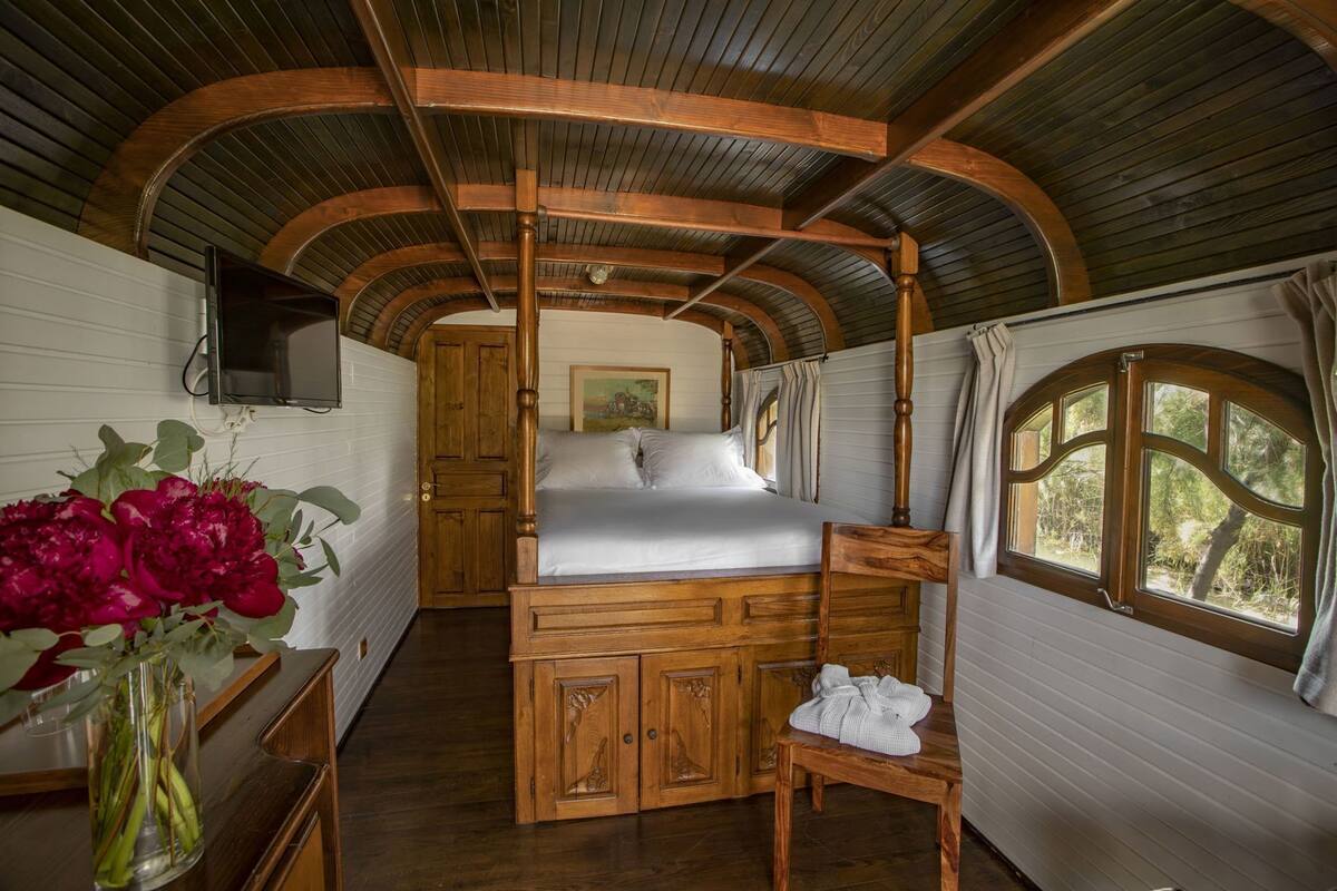 A room with a wooden bed, a wooden chair, a wooden desk, a vase with flowers, and a painting on the wall.