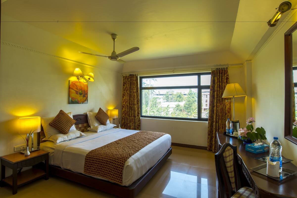 A hotel room with a large bed, bedside lamps, a ceiling fan, a window with curtains, and a desk with a water bottle and tissues.