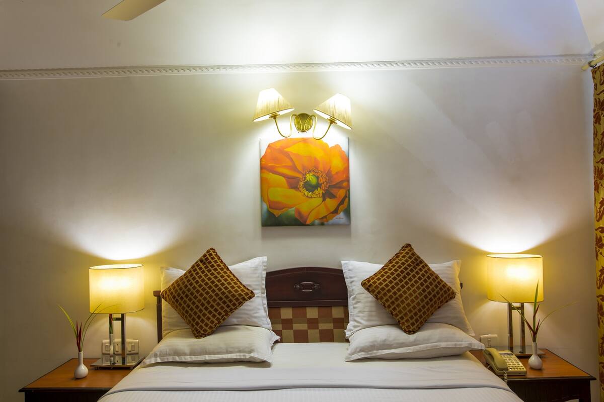 A hotel room with a bed, two bedside tables with lamps, a painting of a flower on the wall, and a checkered headboard.