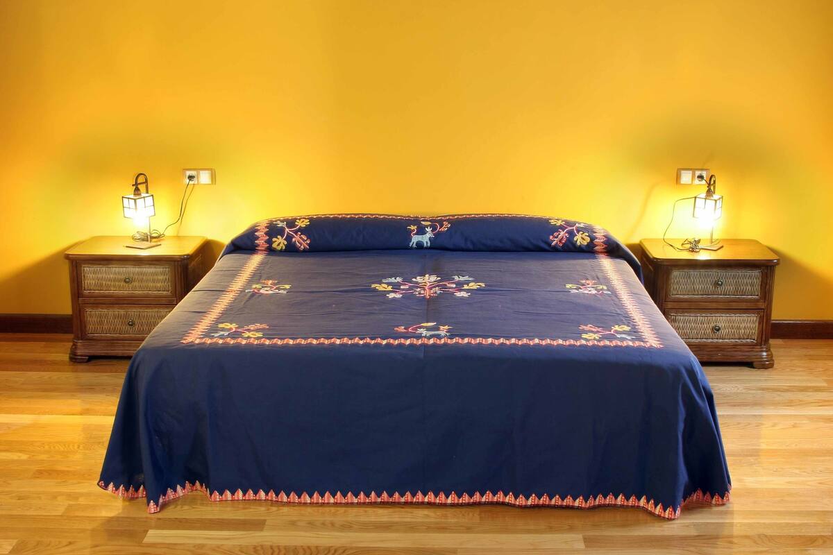Ein Schlafzimmer mit Holzbalkendecke, zwei Nachttische mit Lampen, einem Bett mit Blumenmuster-Überwurf und einer gelben Wand.