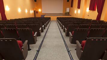 A theater with rows of black seats and red cushions, a stage with a white screen, and red curtains.