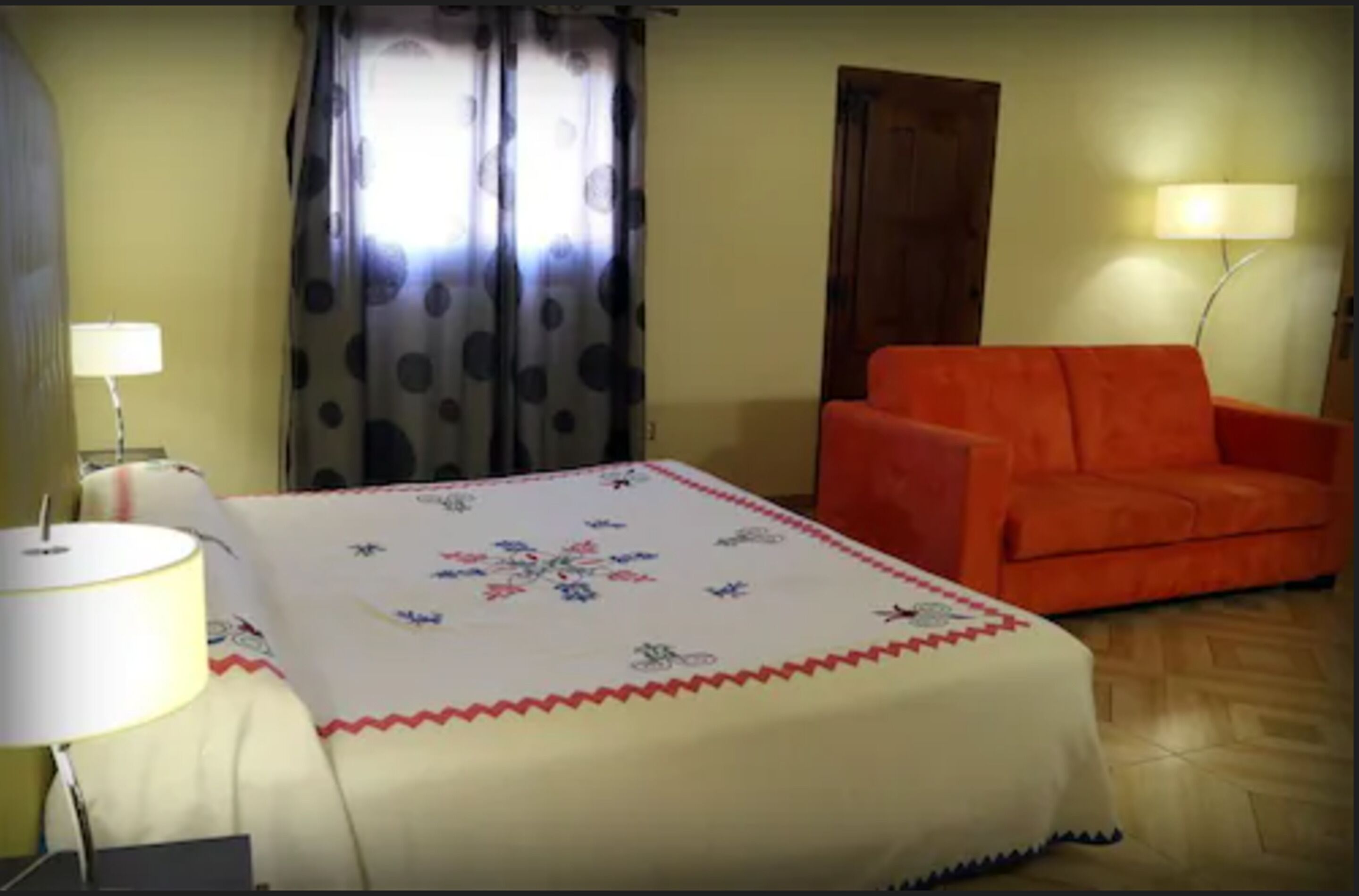 A room with a bed covered in a floral-patterned blanket, an orange sofa, a lamp, and a curtain with a polka dot design.