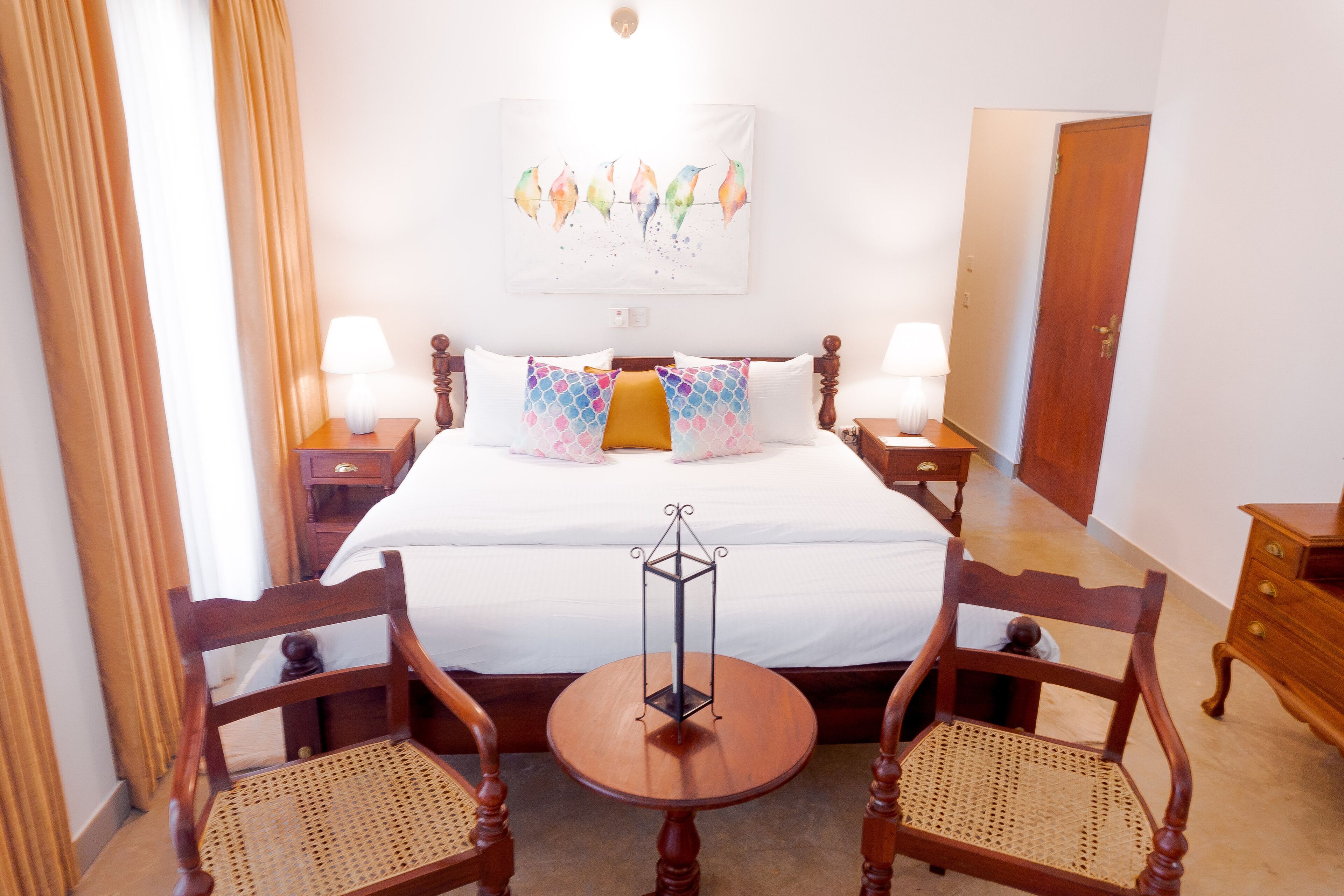 Une chambre d’hôtel avec un grand lit, deux tables de chevet avec des lampes, une petite table ronde, deux chaises en bois et une œuvre d’art accrochée au mur.