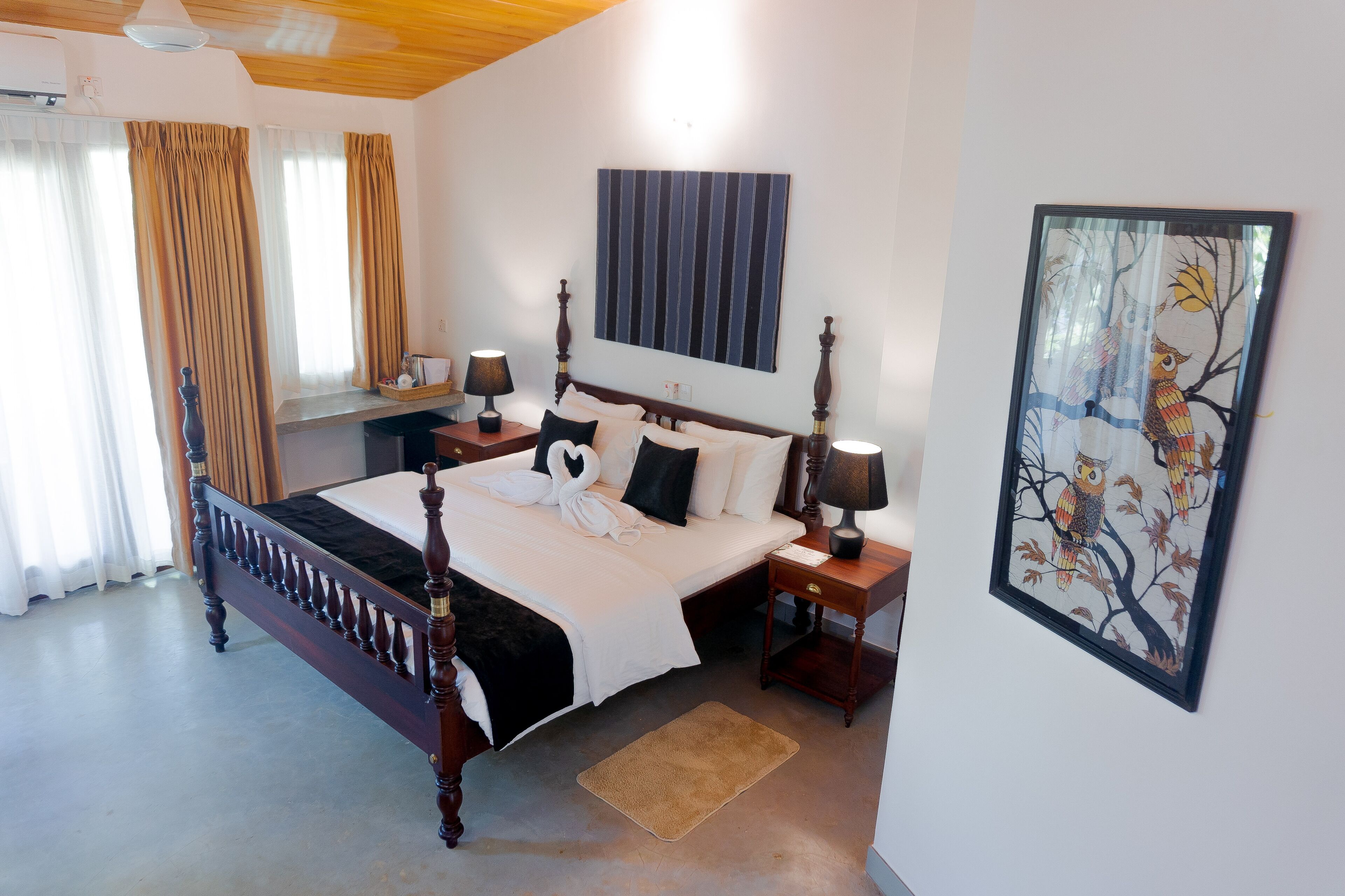 A bedroom with a four-poster bed, a nightstand with a lamp, a framed picture on the wall, and a window with curtains.