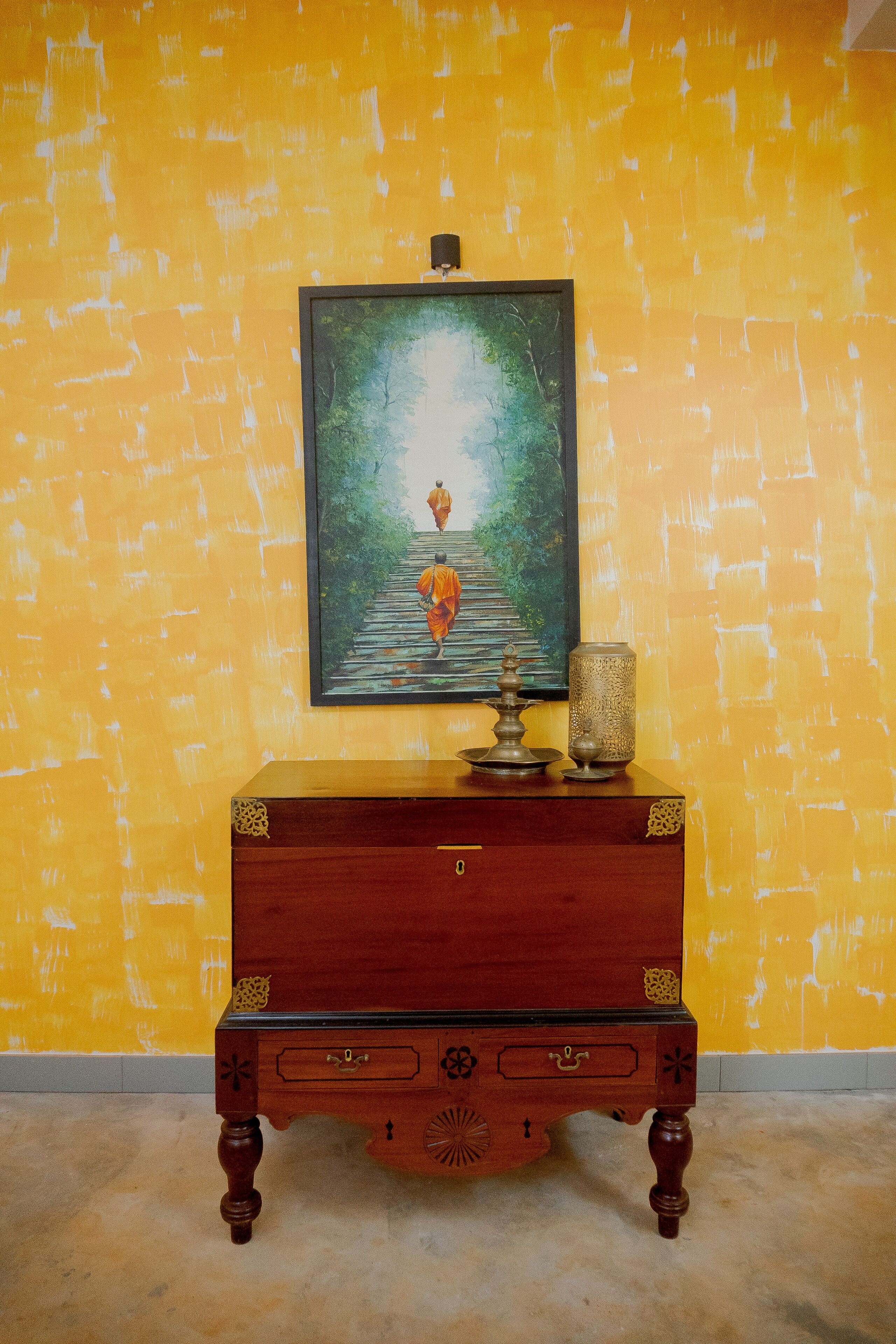 A wooden cabinet with drawers and a decorative item on top, set against a yellow wall with a painting above it.