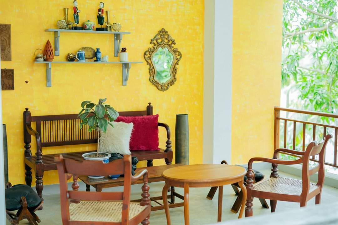 A room with a yellow wall, a wooden bench, two wooden chairs, a small round table, and a potted plant.
