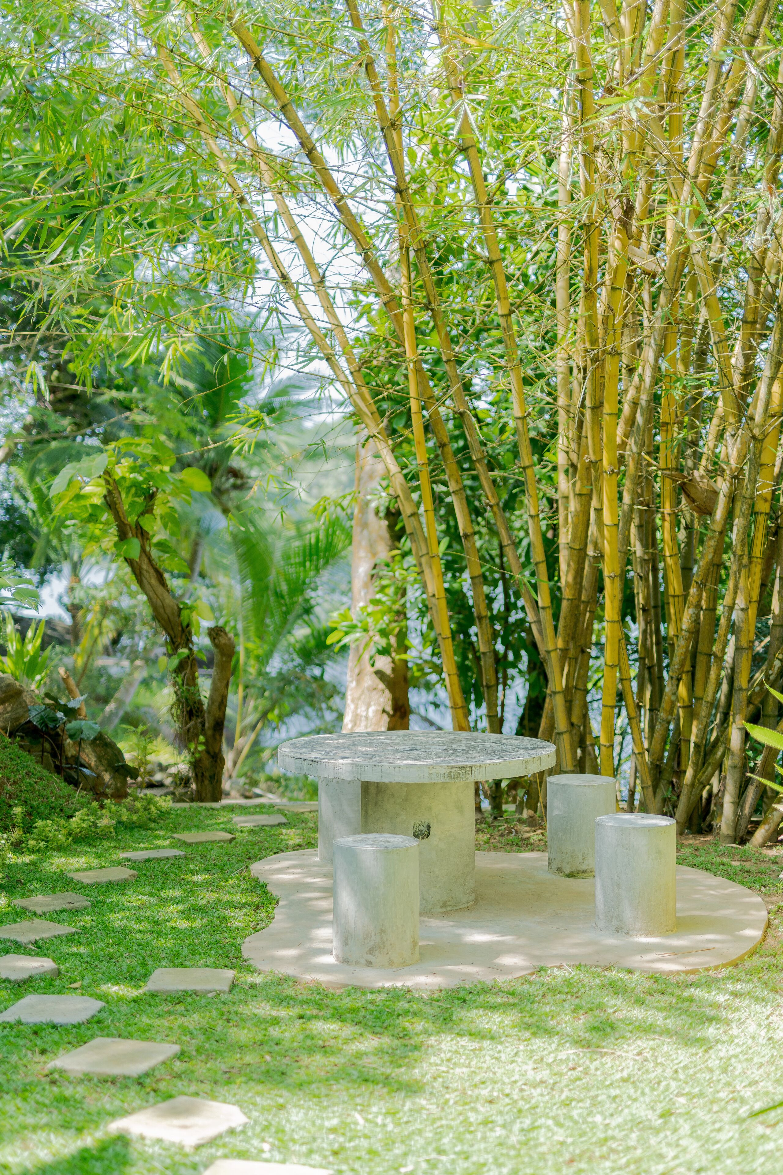 Un jardín con una mesa de piedra circular rodeada de taburetes cilíndricos, ubicada sobre un sendero de concreto con piedras.