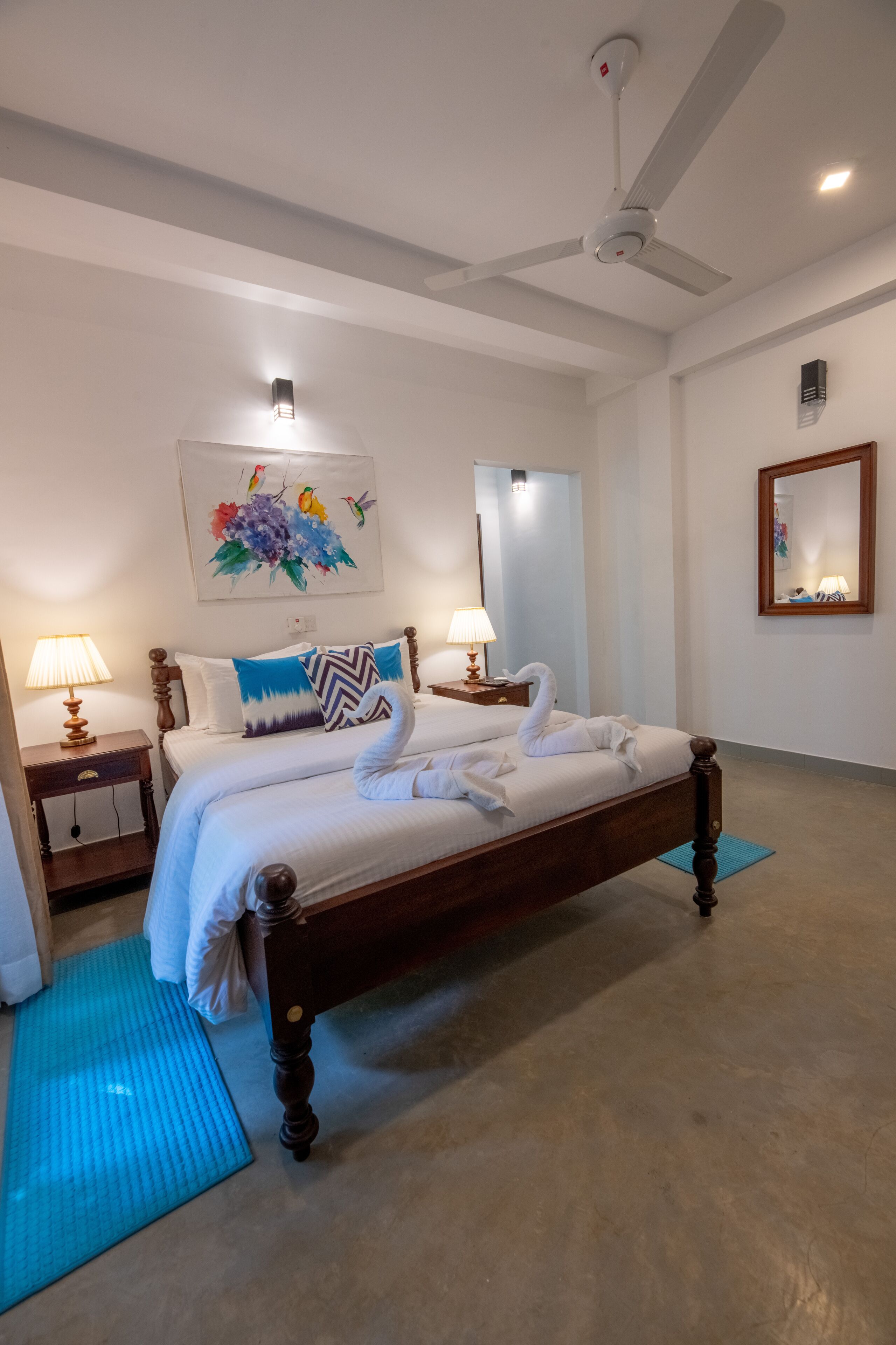 A neatly made bed with a decorative swan pillow and a blue and white patterned pillow. There is a ceiling fan above the bed, and a painting of birds on the wall.