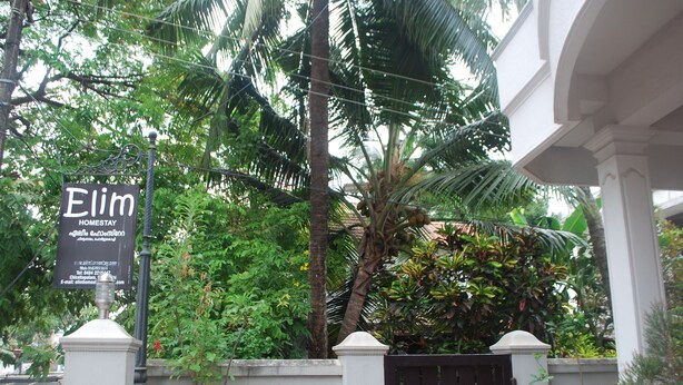 A sign for 'Elim Homestay' with contact information and a view of a white building with a gated entrance, surrounded by lush greenery.