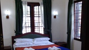 A neatly made bed with white and red bedding, a ceiling fan, and an air conditioning unit. There are two wooden chairs and a small table with a lamp.