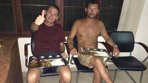 Two men seated on chairs, each holding a tray with food and drinks.