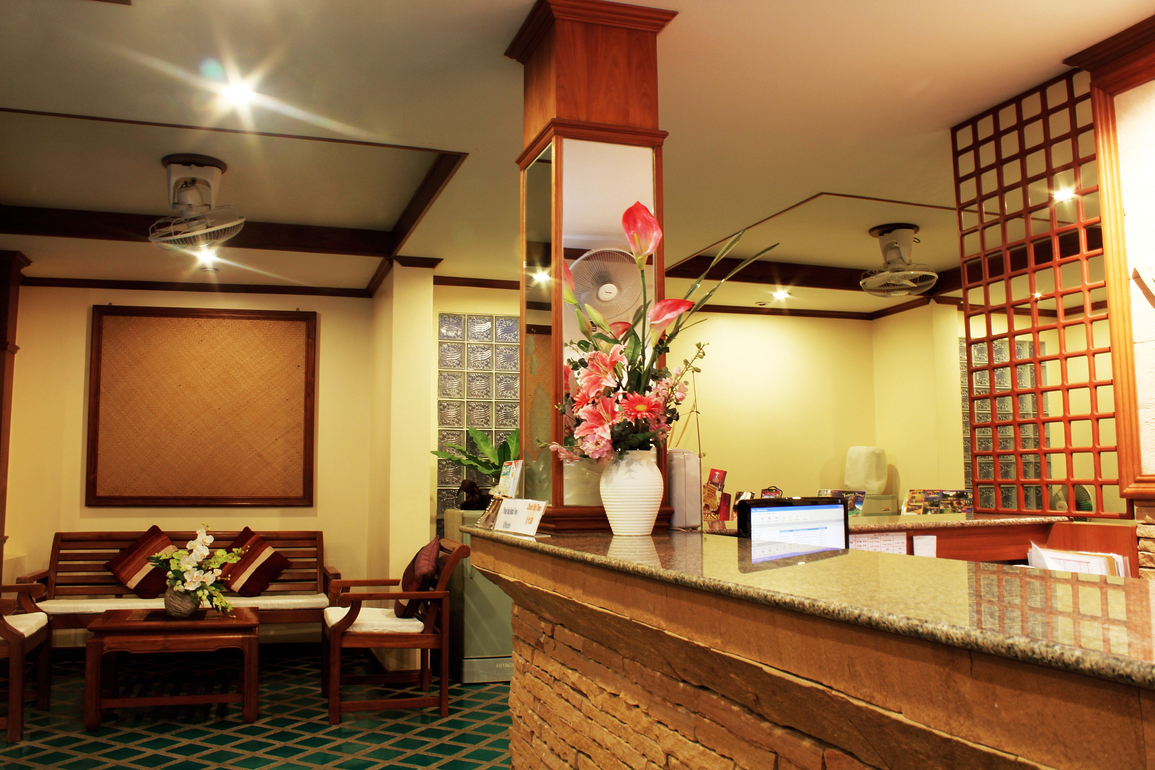 A reception area with a wooden reception desk, a computer, a vase with flowers, a fan, and a seating area with a sofa and chairs.