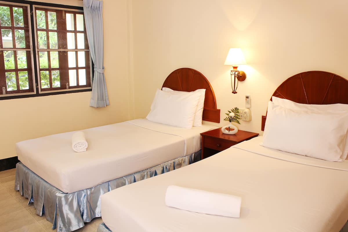 A hotel room with two beds, a nightstand with a lamp, a window with curtains, and a view of greenery outside.