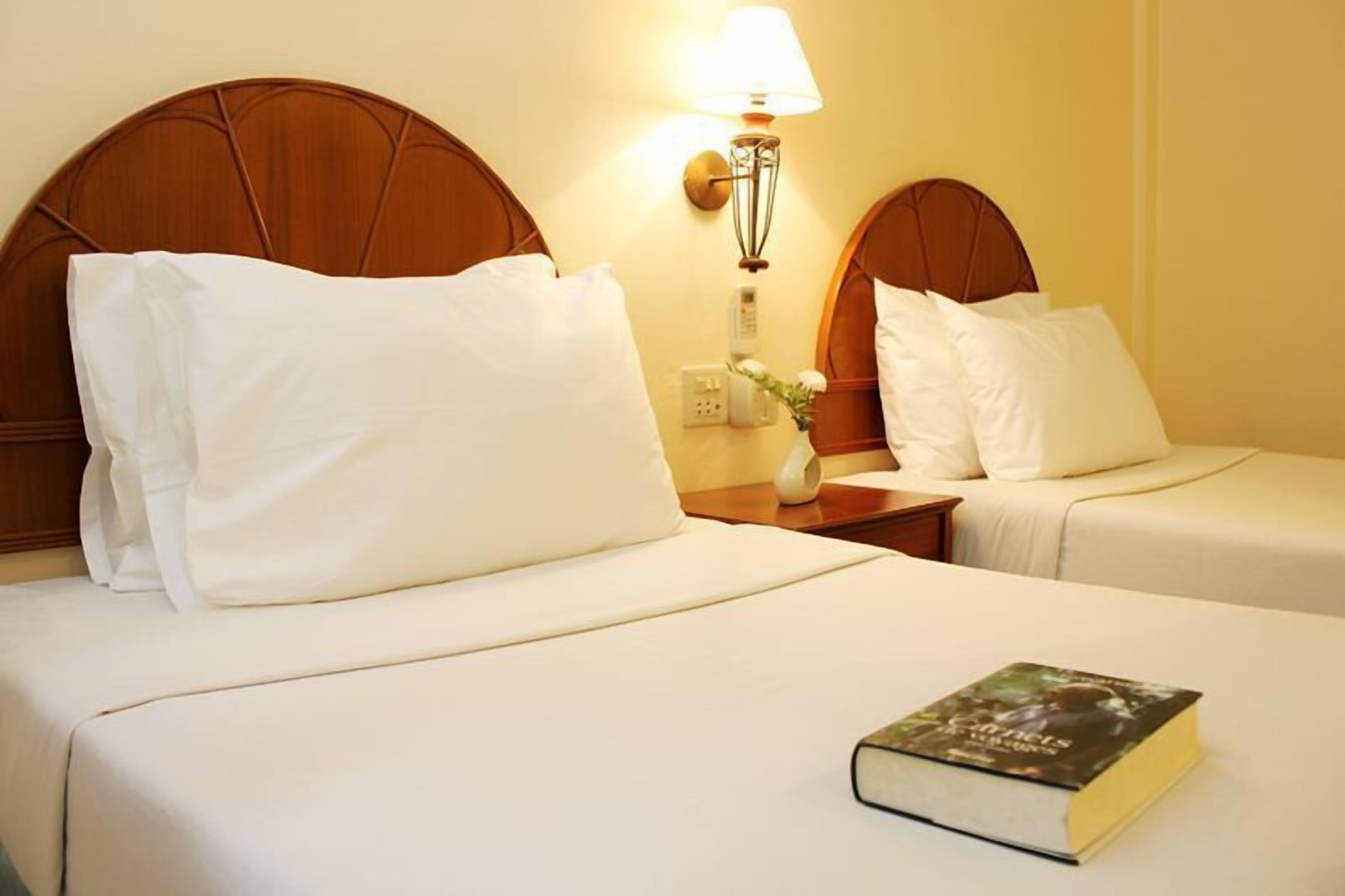 A hotel room with two beds, each with a wooden headboard, white bedding, and a bedside lamp. A book is placed on the bed in the foreground.