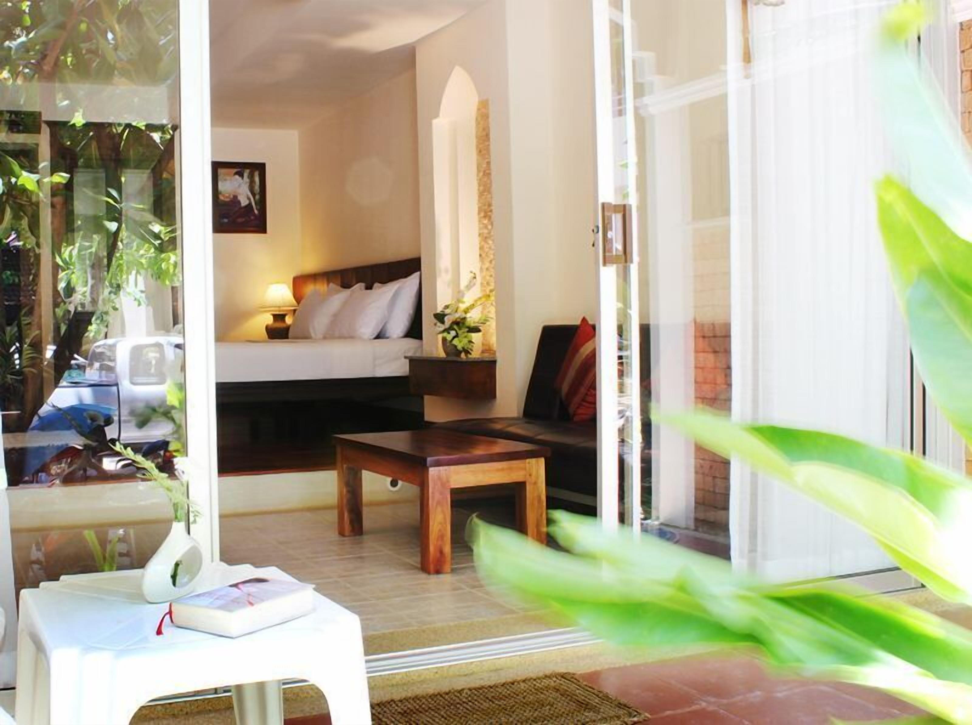 A cozy room with a bed, a small table, and a sofa. There is a window with a view of greenery outside.