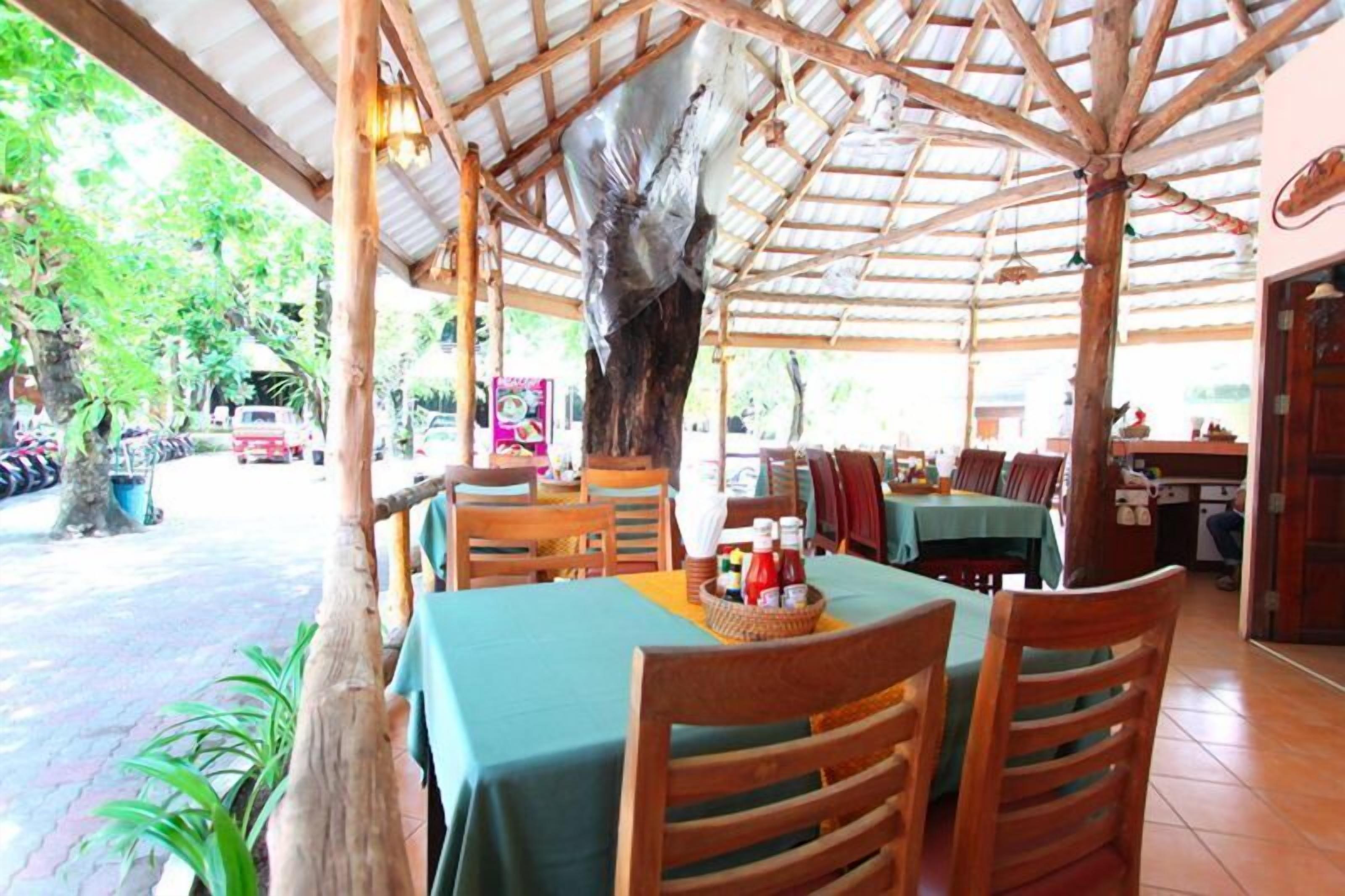 Un restaurant en plein air avec des tables et des chaises en bois, un toit abrité et une vue sur la rue et la verdure.