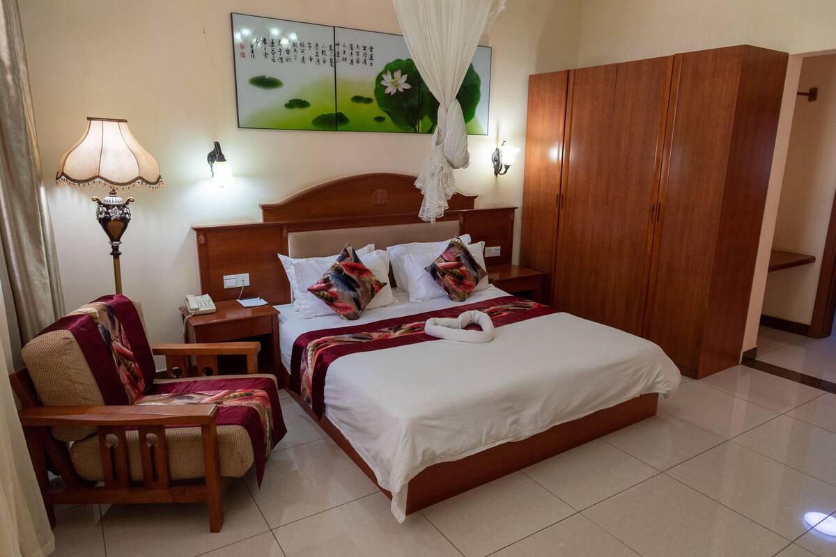 Quarto de hotel com uma cama, um guarda-roupa de madeira, uma cadeira, mesa de cabeceira, abajur e uma obra de arte na parede.