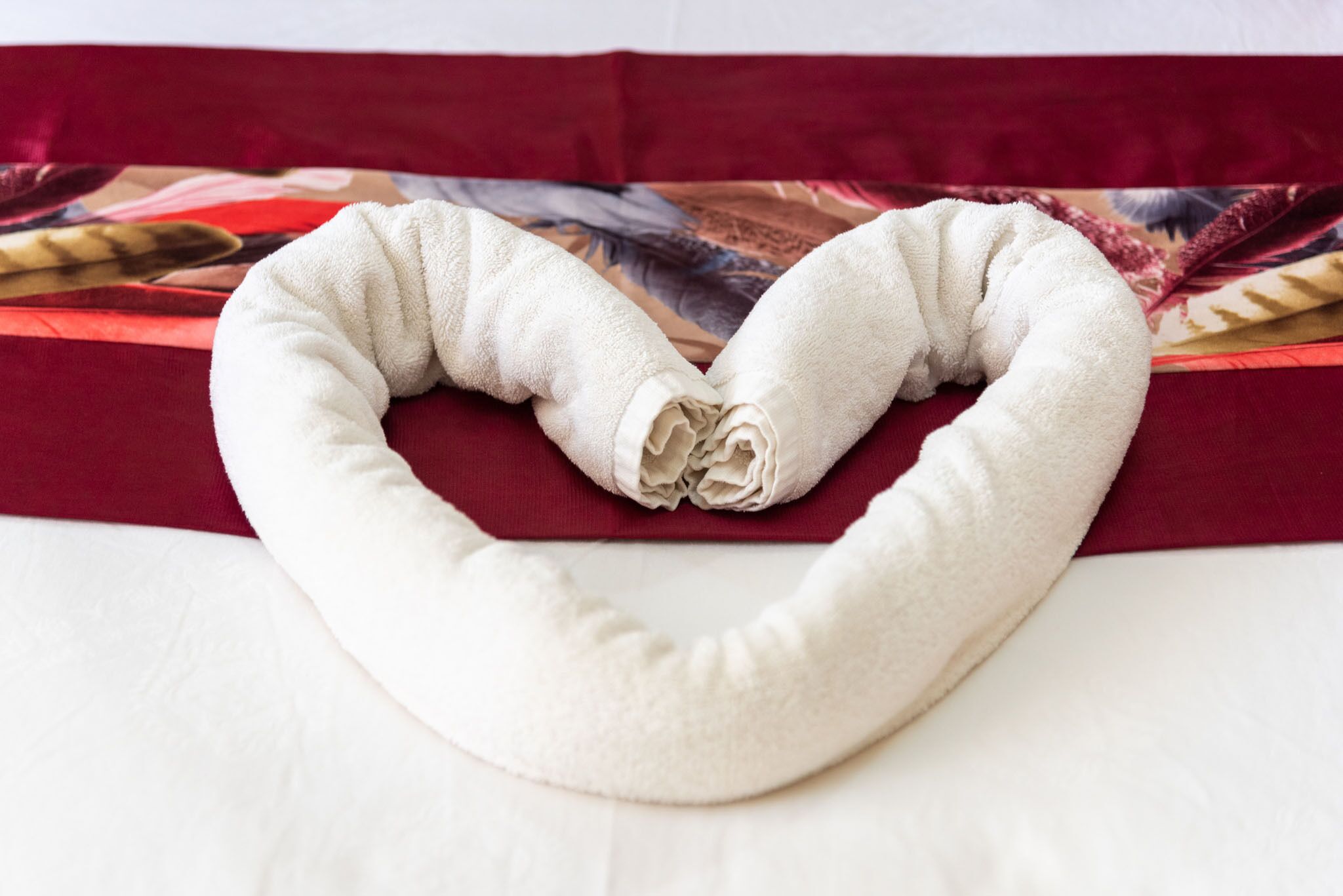 Een wit reiskussen in de vorm van een hart, geplaatst op een bed met een kussenhoes met een rood en beige patroon.