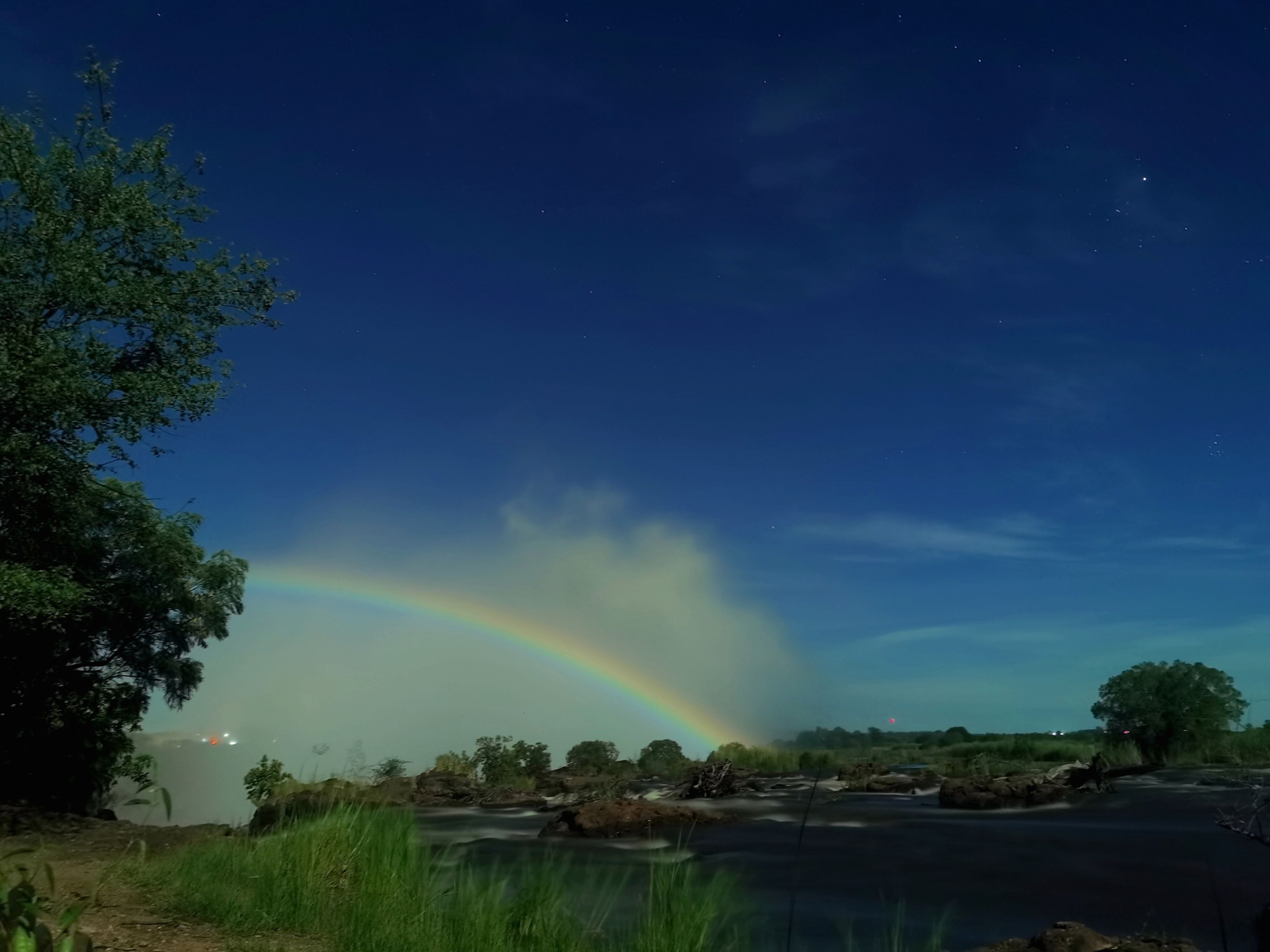 Nachtsäule mit einem deutlich sichtbaren Regenbogen über einem Fluss, umgeben von Bäumen und einem klaren Himmel.