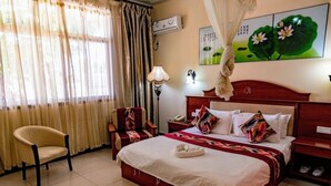 A hotel room with a bed, a chair, a bedside table, a lamp, and a wall-mounted air conditioner. - Oriental Swan Hotel Livingstone (Livingstone)