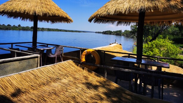 A thatched-roof outdoor seating area with tables and chairs overlooking a body of water. - Oriental Swan Hotel Livingstone (Livingstone)