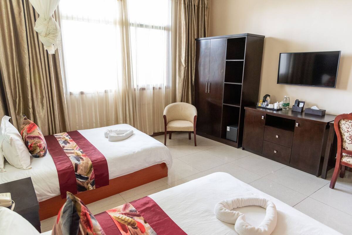 Quarto de hotel com duas camas, uma escrivaninha, um guarda-roupa, uma televisão e uma cadeira.