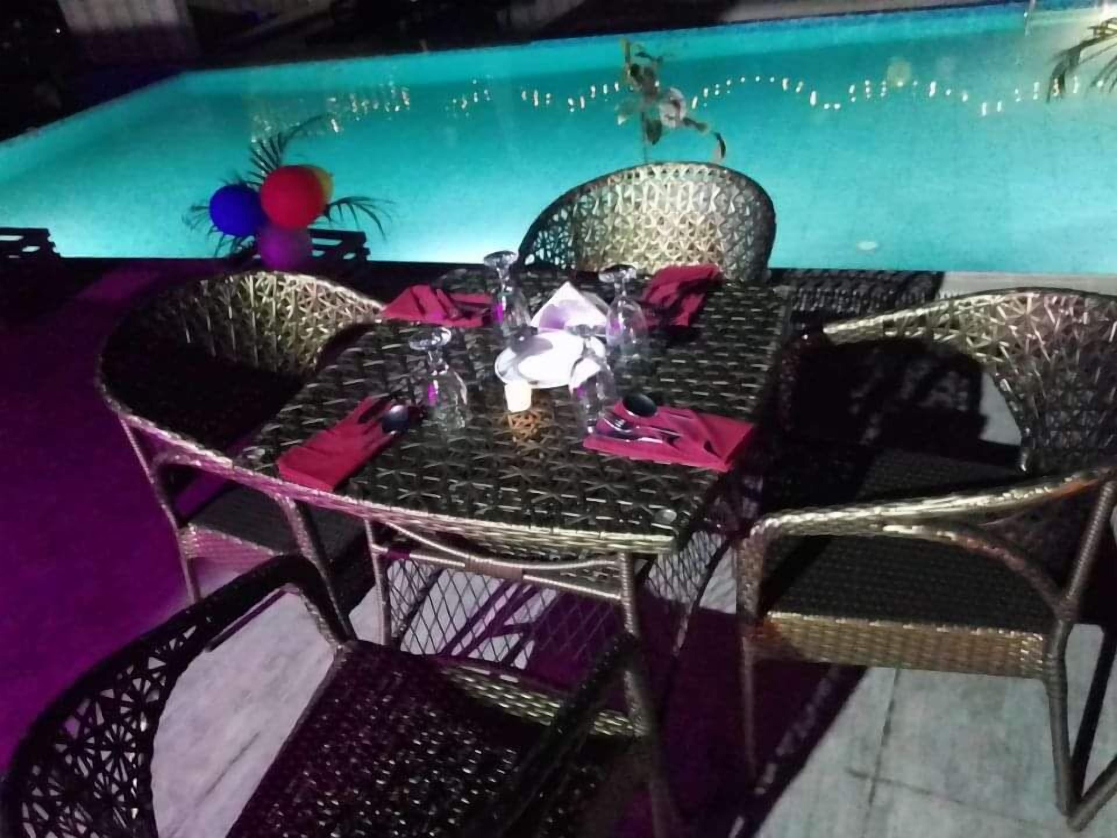 A poolside dining area with wicker chairs and a table, a pool in the background, and a decorative plant.