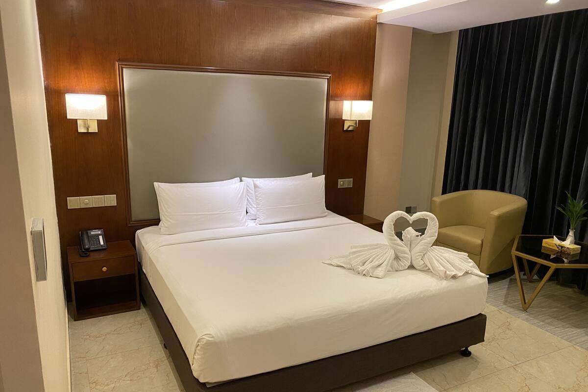 A hotel room with a large bed, a bedside table with a phone, a chair, and a small table with a plant.