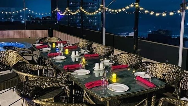An outdoor dining area with tables set for two, surrounded by wicker chairs, under large umbrellas, with string lights and a city view in the background. - Hotel Tower Inn (Chittagong)