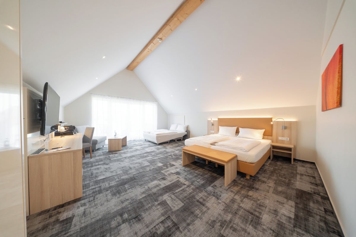A modern hotel room with a sloped ceiling, a large bed, a desk with a computer, a sofa, and a TV.