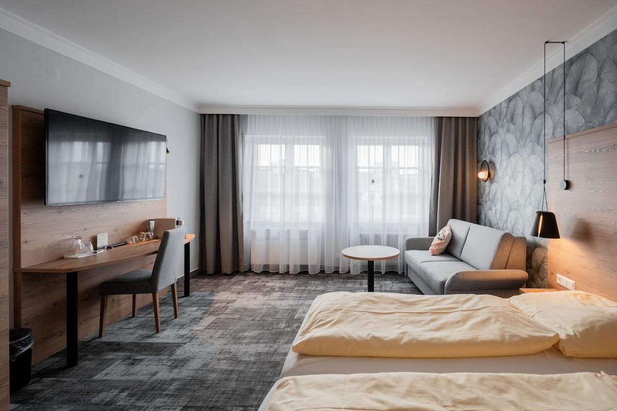A hotel room with a bed, a sofa, a desk with a chair, a TV, and a small table.