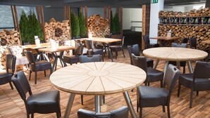 Un restaurant avec des tables rondes en bois et des chaises noires, entouré d’un plancher en bois et d’une pile de bois de chauffage.