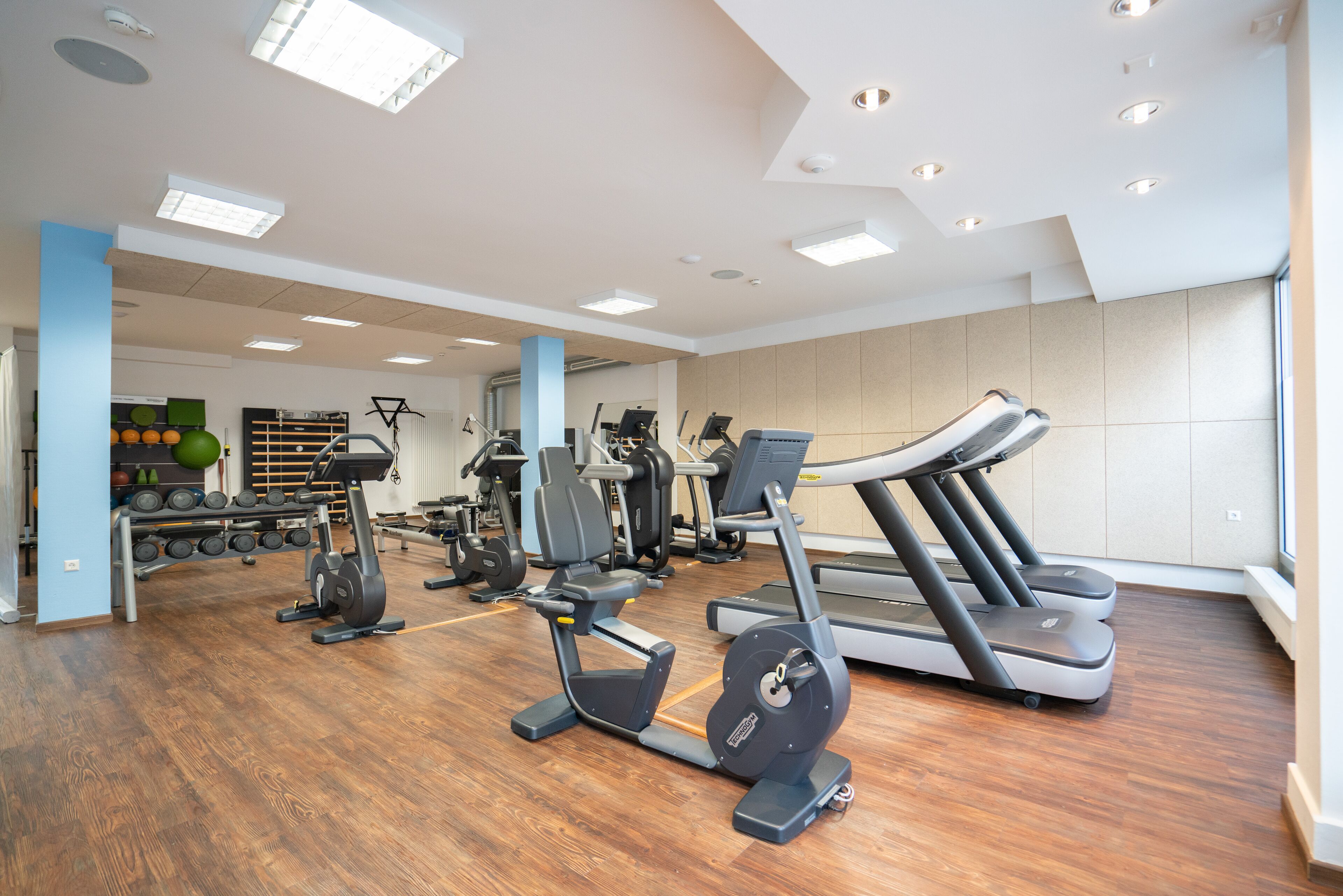 Un gimnasio moderno con diverso equipo de ejercicio, incluyendo cintas de correr y máquinas de pesas, y suelo de madera.
