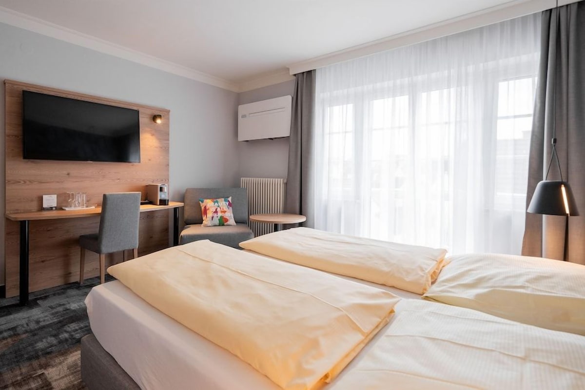 A hotel room with two beds, a desk, a chair, a TV mounted on the wall, and a window with curtains.