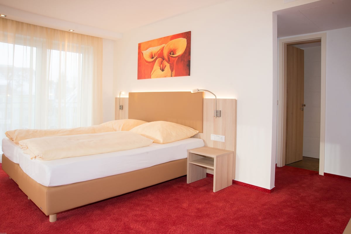 A hotel room with a bed, a nightstand, a wall-mounted lamp, a painting, and a red carpet.