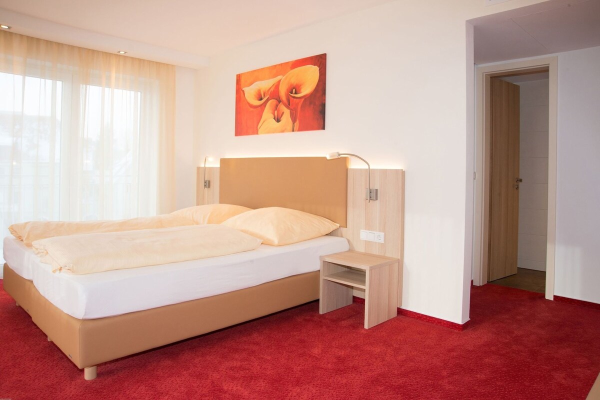 A hotel room with a large bed, a bedside table, a nightstand, a painting on the wall, and a door leading to another room.